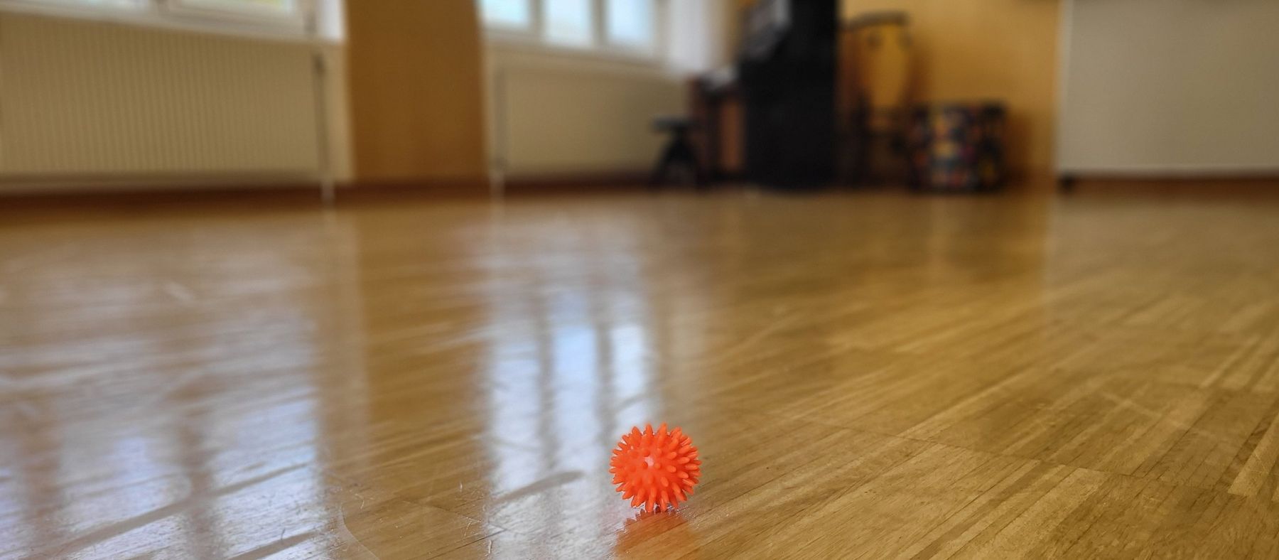Ein oranger Igelball auf dem Parkettboden eines leeren Musikraums, im Hintergrund Klavier und Trommeln